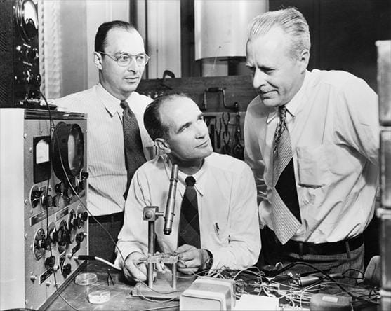 Título: John Bardeen, William Shockley e Walter Brattain em uma fotografia promocional de 1948 dos Bell Telephone Laboratories - Descrição: Fotografia em preto e branco de John Bardeen, William Shockley e Walter Brattain reunidos ao redor de equipamento eletrônico.